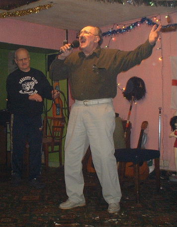 Gordon with right hand holding a microphone to his wide open mouth and the left flung open in extravagant gesture apparently singinging impassionately, while behind Oz starts coiling up the unplugged microphone lead.